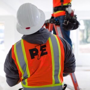 An electrical inspector and worker wearing hard hats and reflective vests are working indoors; one stands on a ladder, and the other faces away, featuring the initials "PE" on their vest.