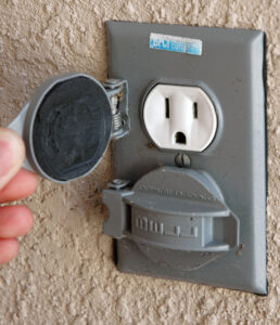 A hand opens an outdoor electrical outlet cover mounted on a textured wall.