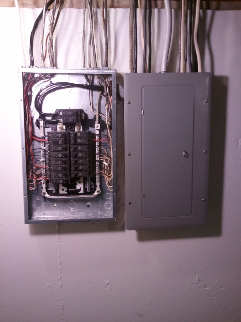 Open electrical panel with multiple circuit breakers and wires next to a closed panel door, mounted on a wall.