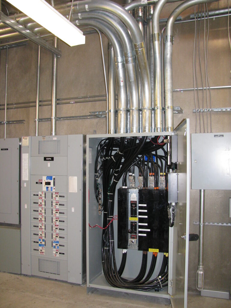 Open electrical panel with multiple cables and switches, surrounded by conduit pipes against a gray wall.