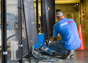 A technician in a blue shirt with the "PRAIRIElectric" logo kneels on the ground, working on wiring and electrical equipment next to a metal cage and orange traffic cone. As an electrician in Vancouver WA, they ensure every connection is safe and secure.