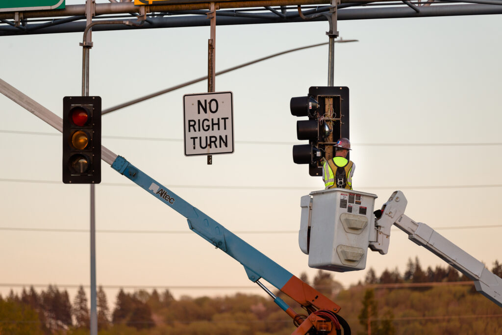 A worker in a lift bucket repairs a traffic signal next to a "No Right Turn" sign, with another lift arm nearby. Trees and power lines are visible in the background.