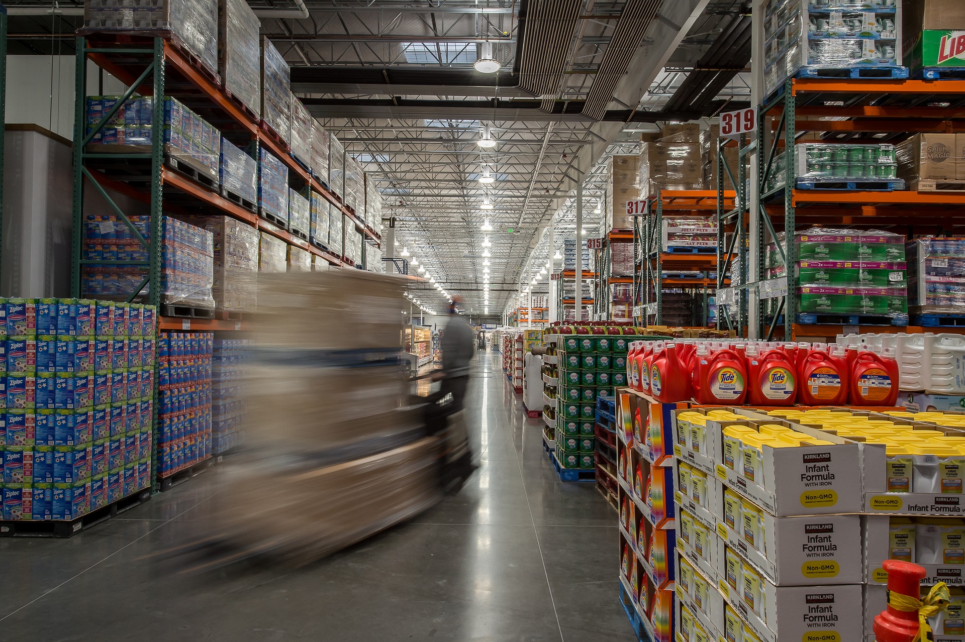 A worker drives a forklift carrying boxes of goods along an aisle in a large warehouse stocked with various products.