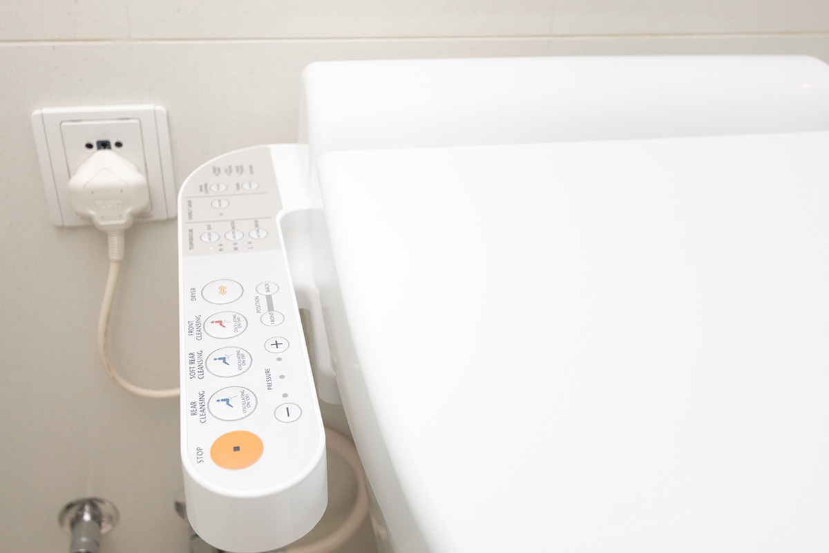 Close-up of a modern toilet with an attached bidet control panel. The panel displays various buttons for different functions beside the toilet seat, with a plugged electrical outlet in the background.