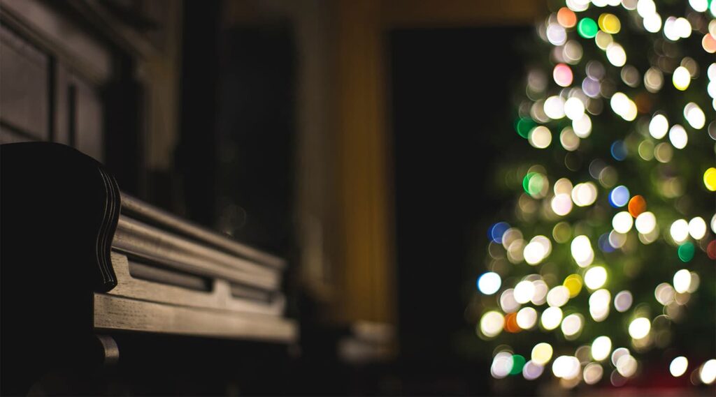 A dark room with a brightly lit, blurred Christmas tree in the background and an elegant wooden piano in the foreground, illuminated softly by electrical lights.