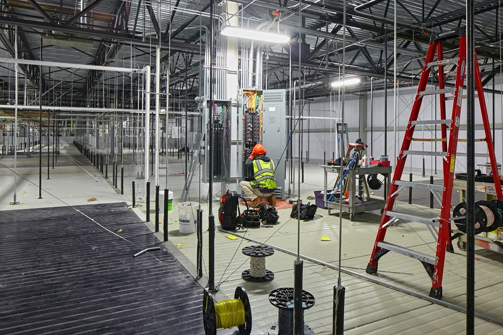 A worker wearing a neon safety vest and hard hat is performing electrical work inside an industrial facility. Nearby are tools, a red ladder, wiring spools, and construction materials.