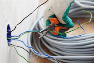 A gloved hand uses wire cutters to trim electrical wires emerging from a wall, expertly managed by an electrician in Vancouver, WA. Coiled cables are in the background.