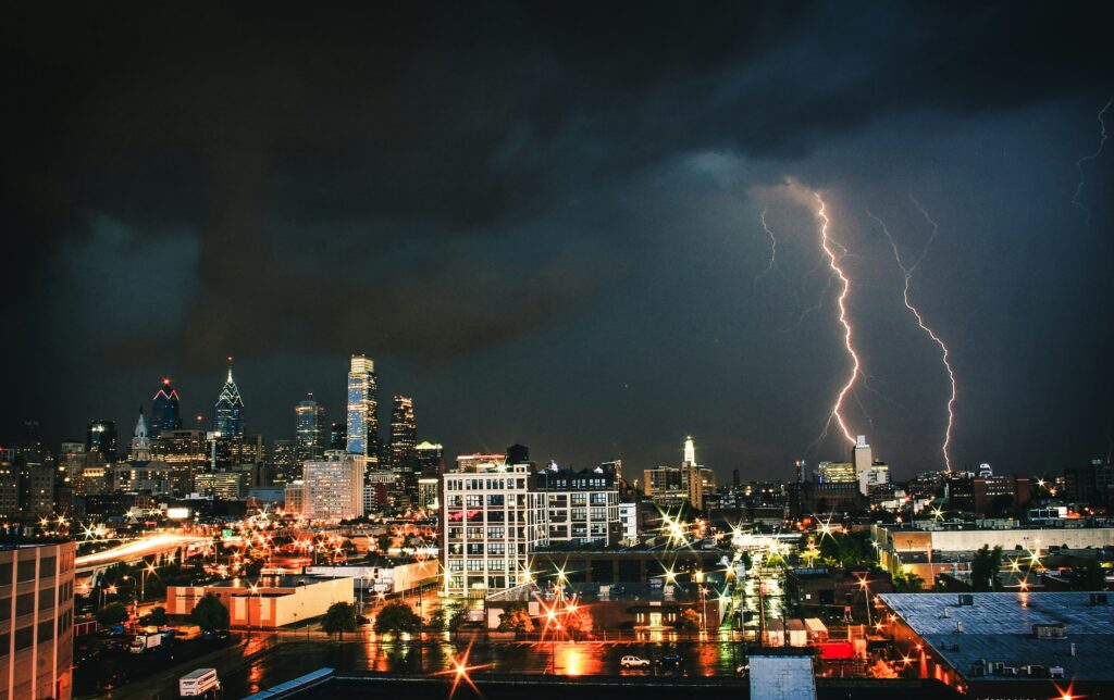 Bright lightning illuminates a cityscape at night, highlighting tall buildings and busy streets below. Dark storm clouds fill the sky, creating a dramatic scene over the urban environment.