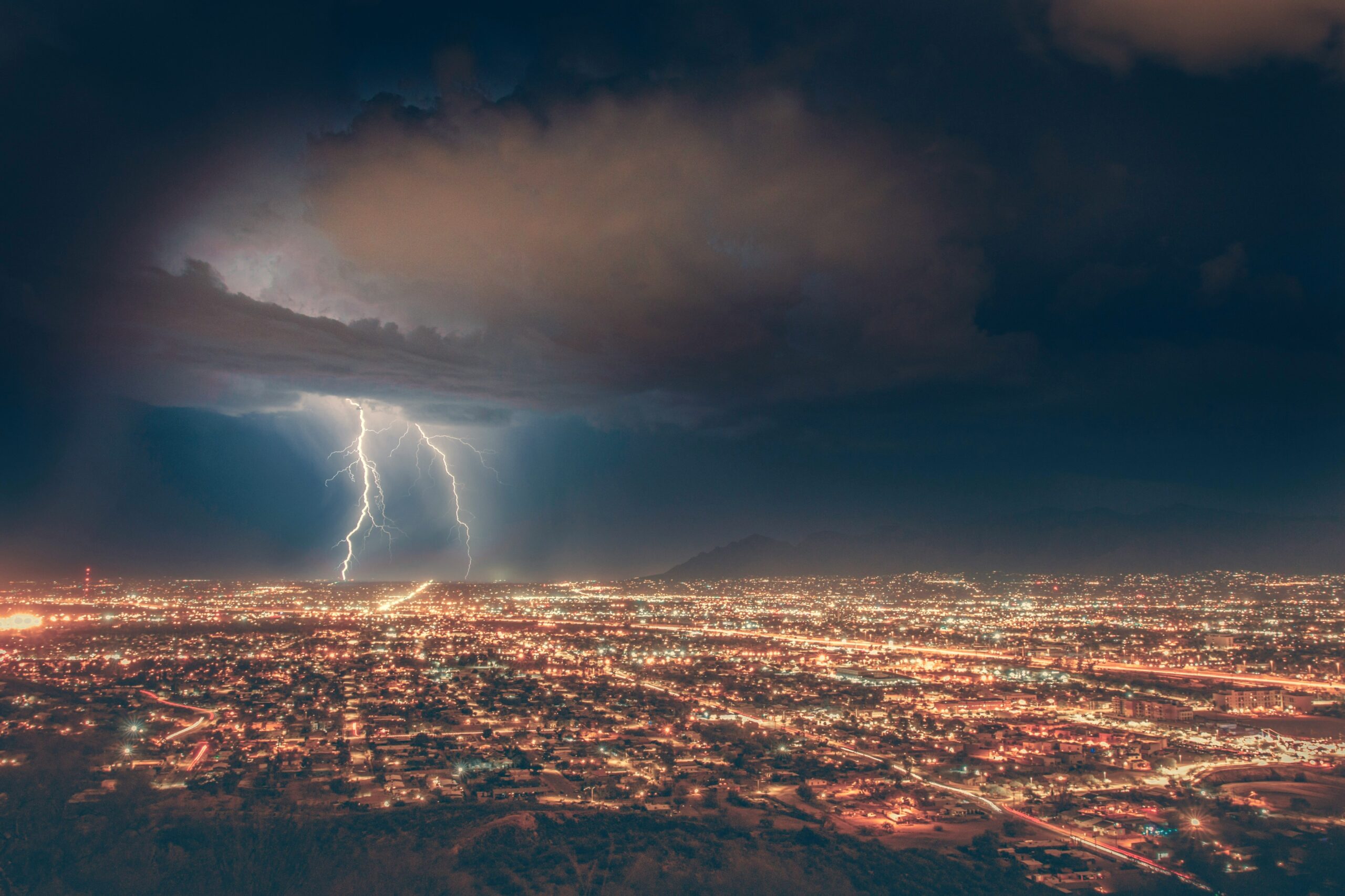 A cityscape at night with a lightning storm in the background, showing bolts of lightning striking through heavy clouds.