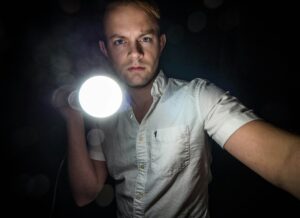 Person holding a bright flashlight, wearing a white button-up shirt, standing against a dark background.