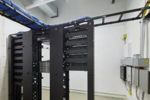 Image of a server room with black server racks, network cables organized along the ceiling, and electrical panels on the wall. The space is clean and has a modern industrial design.