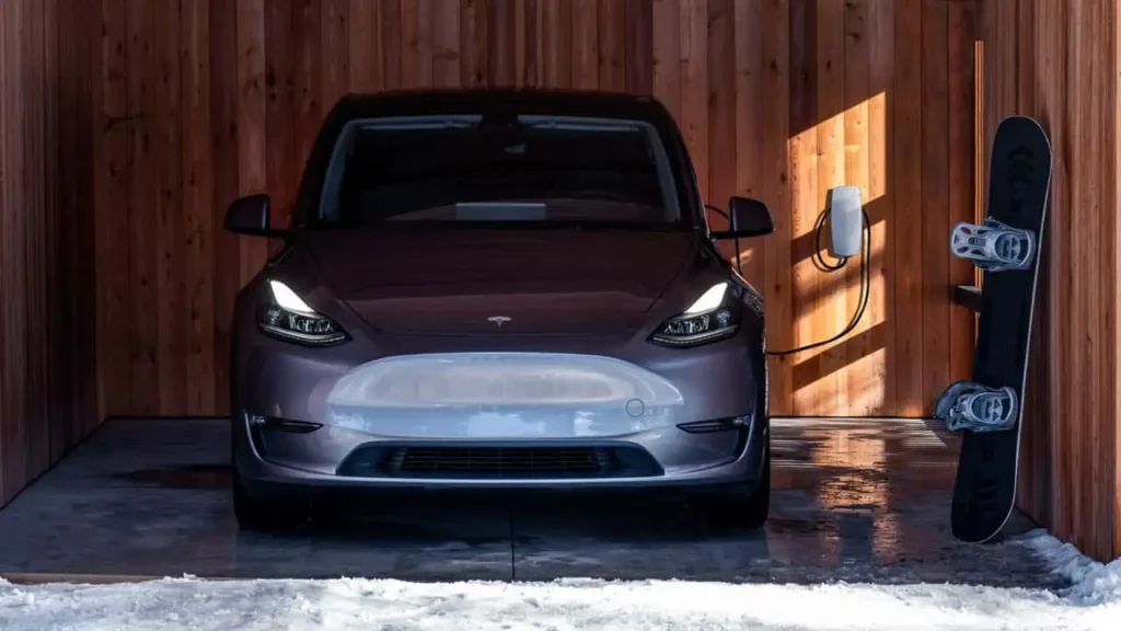 A dark-colored electric car is parked in a wooden garage next to a mounted snowboard.