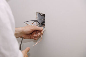 A person is carefully replacing an electrical outlet, handling wires inside a wall-mounted box with one hand holding a wire and the other a tool.